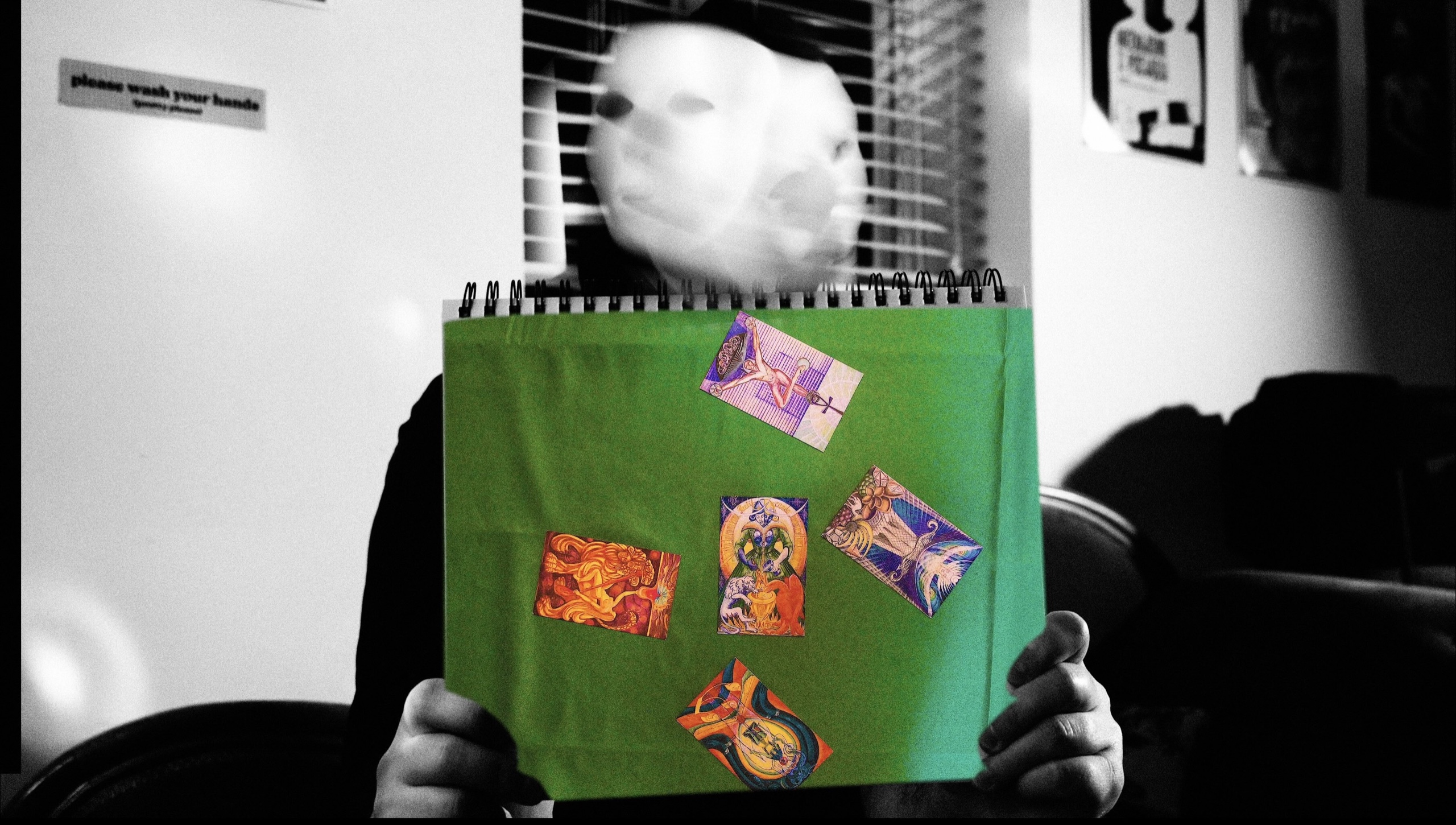 A white-masked figure holding a green notebook covered with colorful tarot cards.