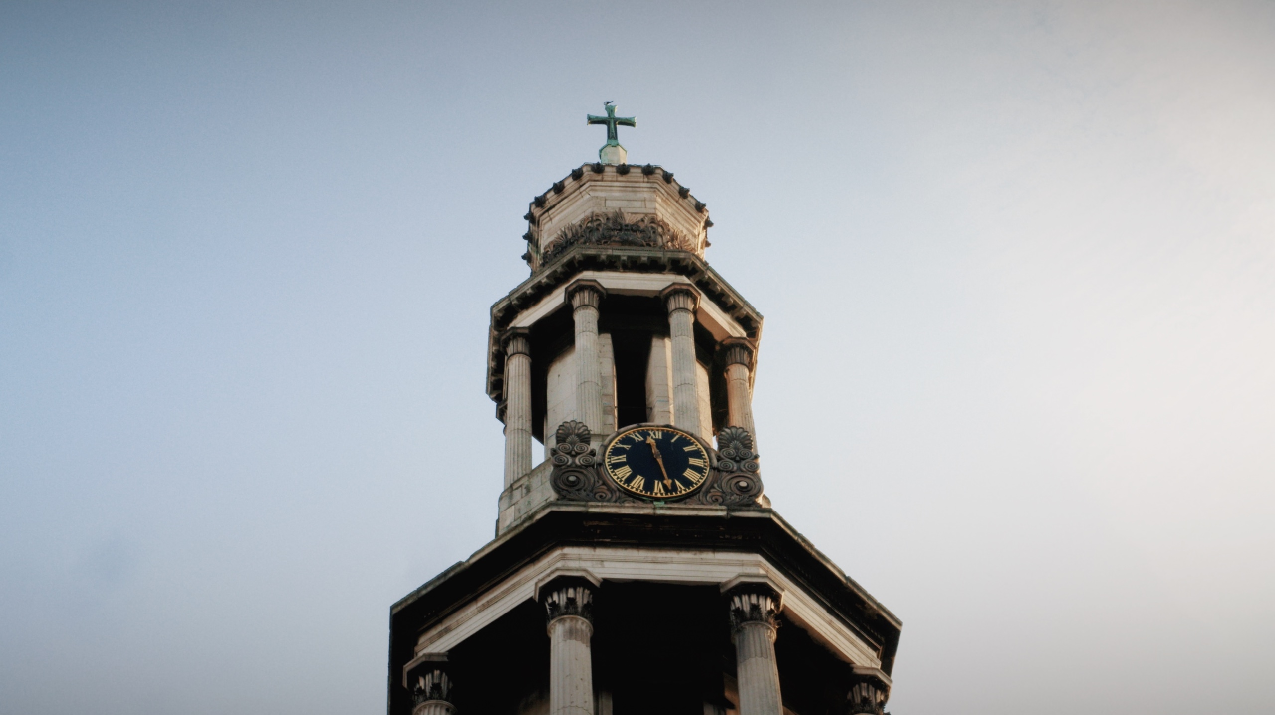A stone church clock tower and cross against a pale blue sky.
