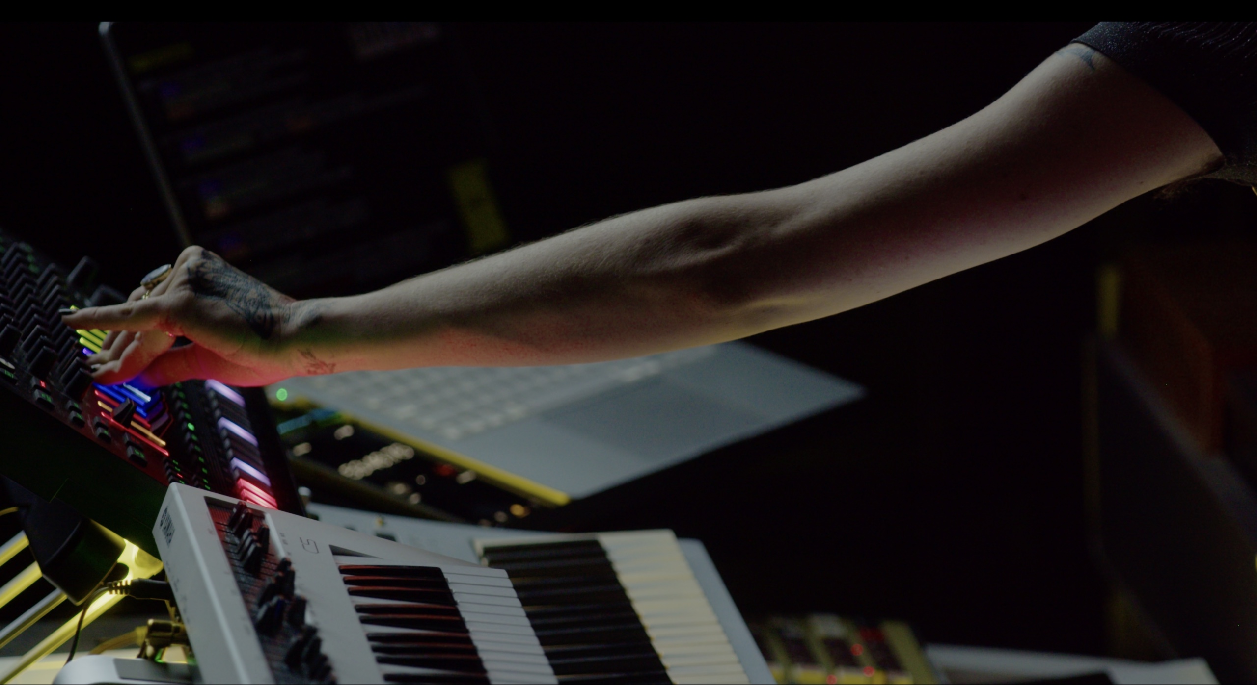 A musician reaching across illuminated synthesizer controls.